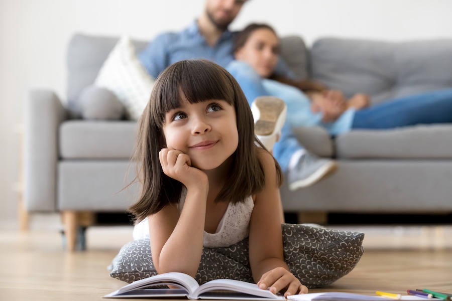 Close up little adorable thoughtful smiling daughter dreaming lying at cushion on warm floor with book in living room at modern home, resting married couple parents on background, focus on small kid.