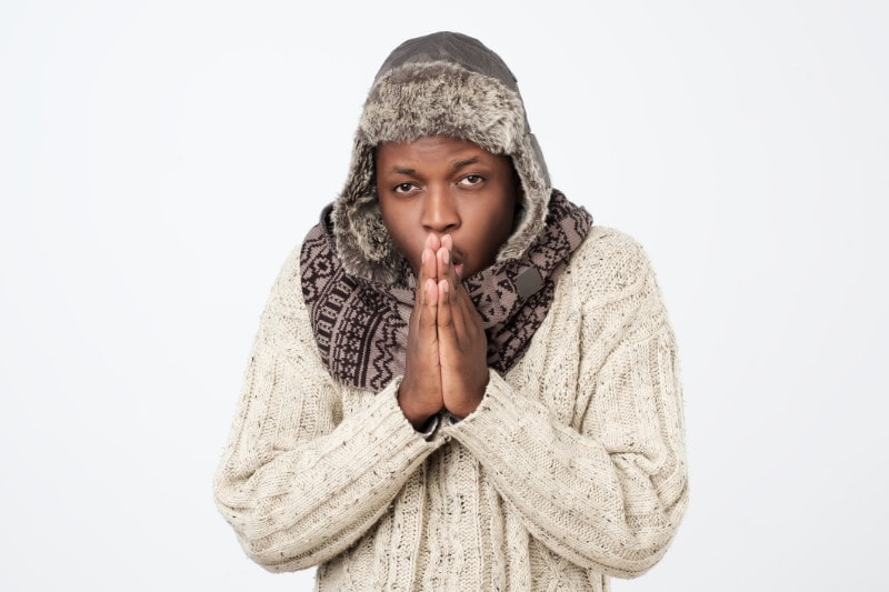 A young black man holding his hands by his mouth to warm up because he is freezing.