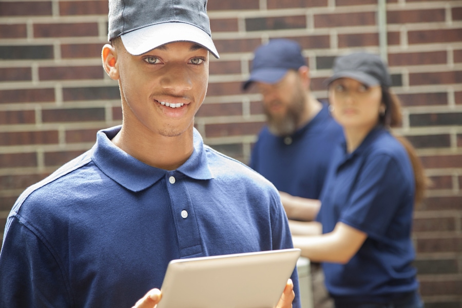 Multi-ethnic group of two men and one woman repairing a home's air conditioner unit outdoors. African descent man in foreground uses a digital tablet to access the repairs. They all wear blue uniforms.