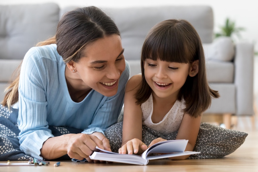 Attractive mother with little adorable daughter lying together at cushions on wooden warm floor in living room at home reading a book fairy tale close up. Weekends activities and development concept.