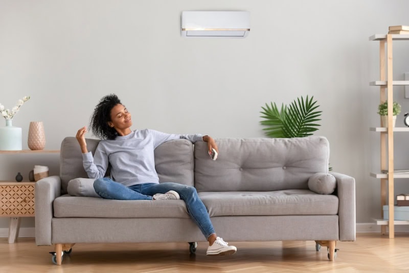 DUC_B_001-woman-under-ductless-ac-1 Ductless Mini Splits for Comfortable and Healthy Living. African american relaxed woman sitting on comfortable couch in living room at modern home holds air conditioner remote control enjoying breathing fresh cool air at summer or warm air at winter season.
