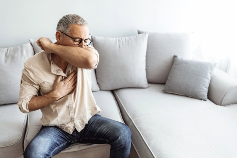 FR_B_002-GettyImages-1306472035 How to Spot a Cracked Heat Exchanger. Man coughs in his elbow while sitting on sofa at home.
